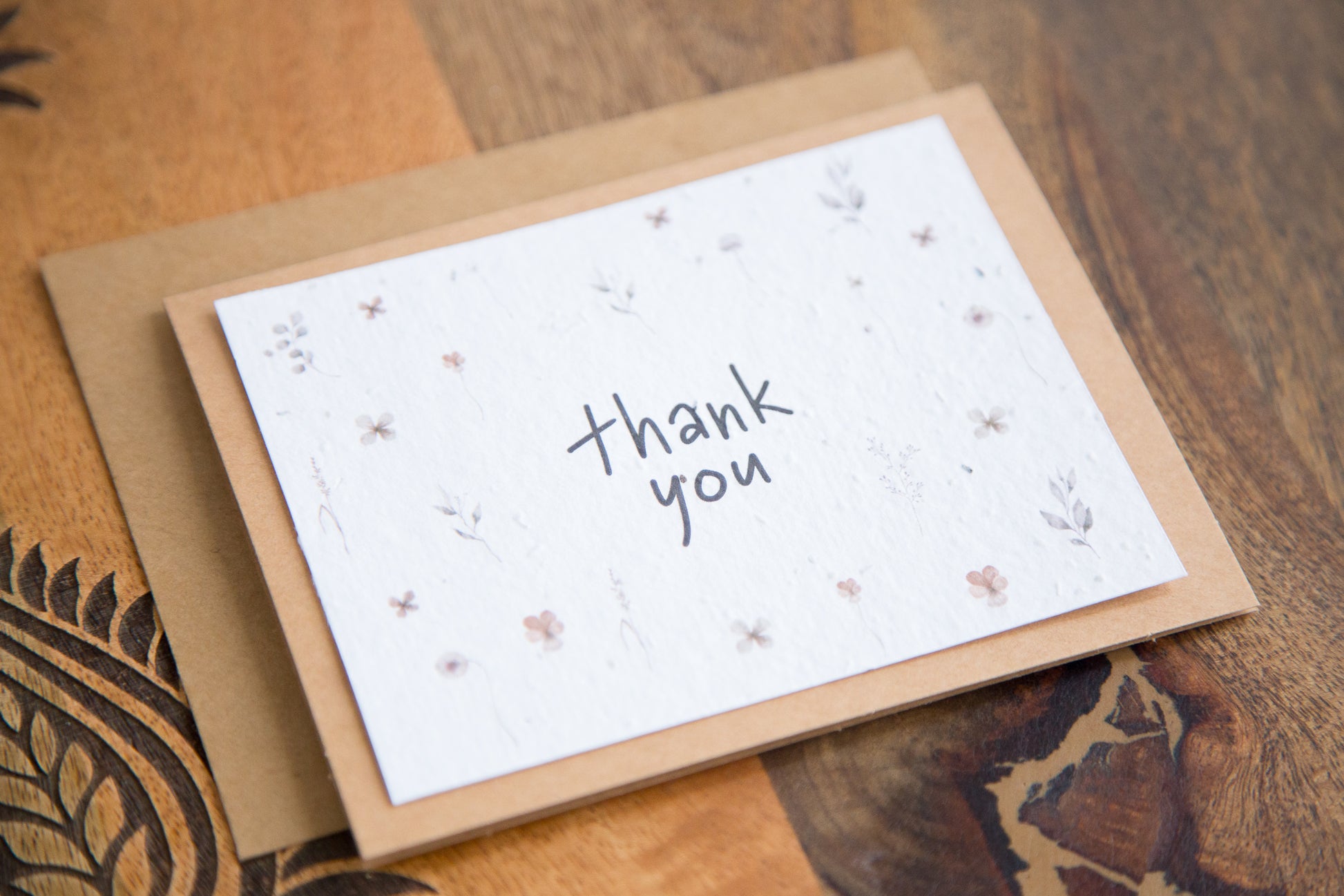 An A2 greeting card on a wooden table with the words "thank you" surrounded by watercolour florals printed on white seed paper 