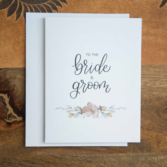 A white A2 sized wedding card with floral designs and the text 'To the bride & groom' on the front, accompanied by a white envelope, displayed on a wooden surface.