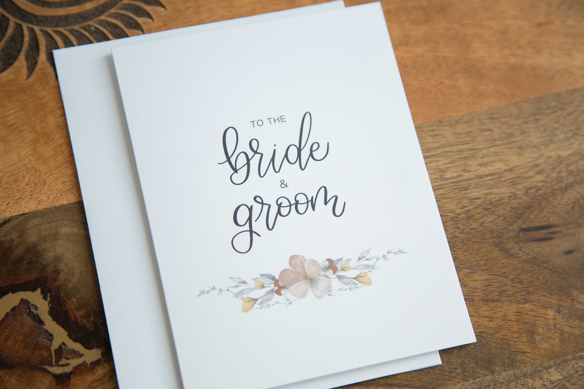 A white A2 sized wedding card with floral designs and the text 'To the bride & groom' on the front, accompanied by a white envelope, displayed on a wooden surface.