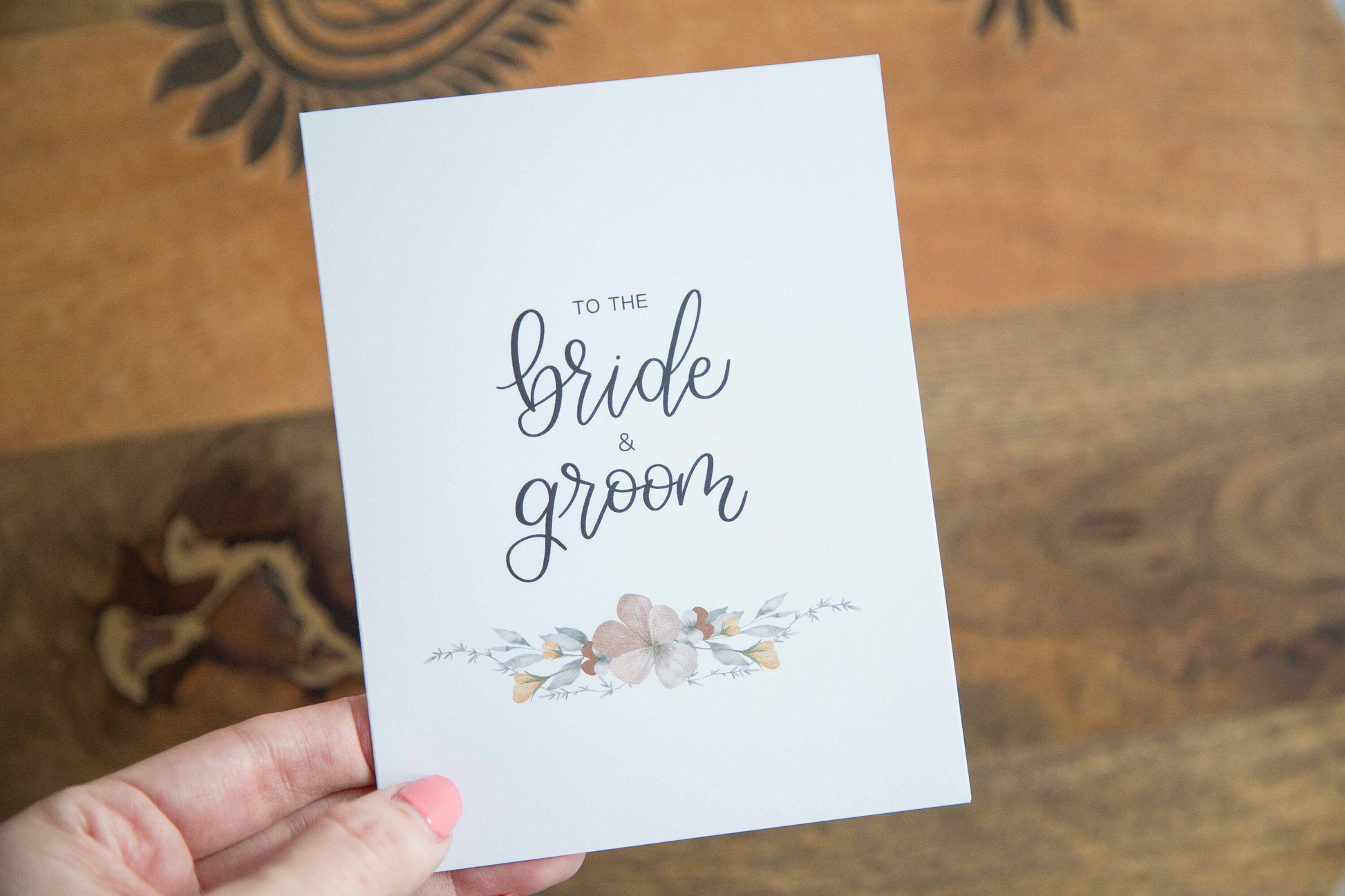 A white A2 sized wedding card with floral designs and the text 'To the bride & groom' on the front, accompanied by a white envelope, displayed on a wooden surface.